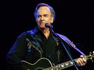Neil Diamond in a black shirt with his guitar behind a microphone